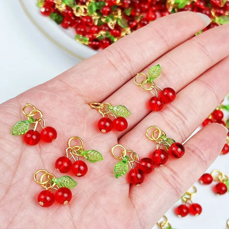 A hand holds several red cherry glass charms with gold-colored wire loops and green leaves.