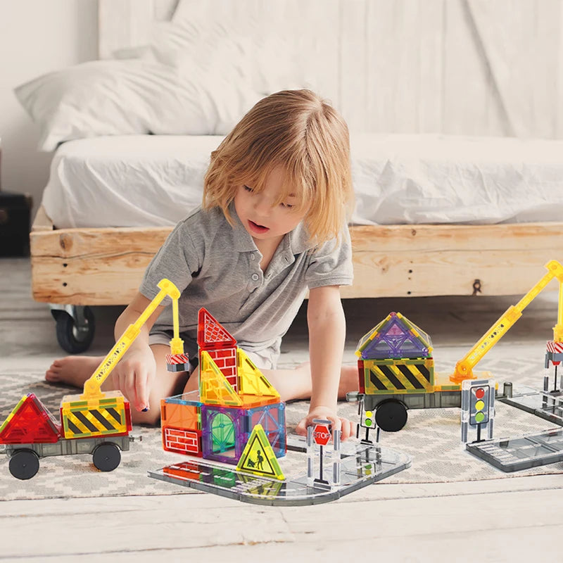 Ein kleines Kind baut ein magnetisches Zugset mit bunten Blöcken und Fahrzeugen auf einem Holzfußboden.