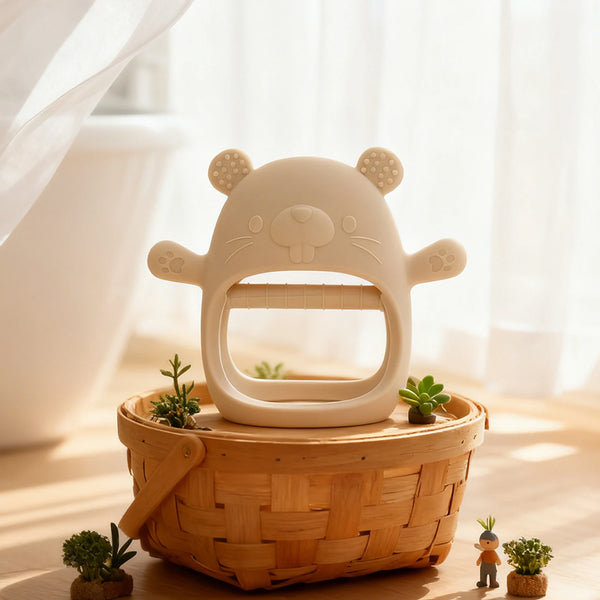 A white silicone teething mitten shaped like a bear sits in a woven basket with small plants.