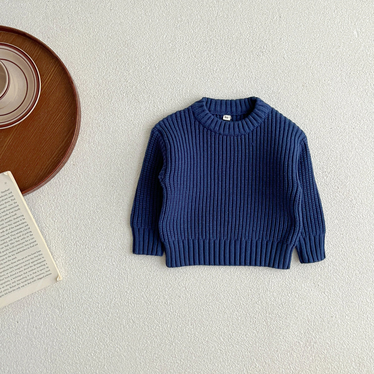 A navy blue knit baby pullover sweater lying flat on a light surface next to a wooden plate and a book.