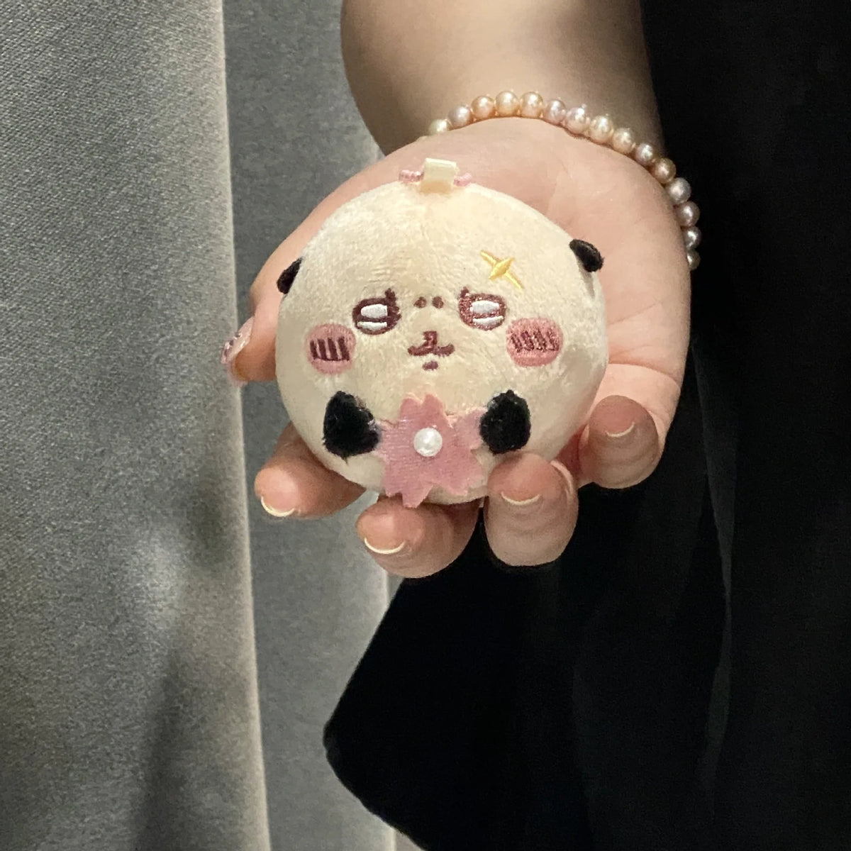 A hand holds a round, light pink plush charm featuring a black face and a pink cherry blossom flower.