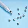 A silver precision bead and jewelry pick-up tool with a textured handle and several small silver beads scattered on a light b