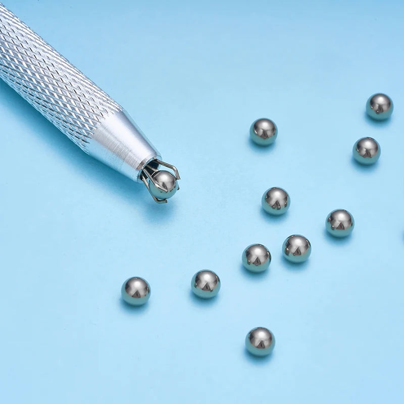 A silver precision bead and jewelry pick-up tool with a textured handle and several small silver beads scattered on a light b