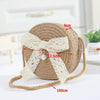A beige handwoven straw crossbody bag featuring a white lace bow and a long tan strap.