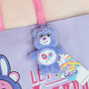 A purple Care Bears plush toy with a light blue face and pink accents is attached to a purple fabric bag.