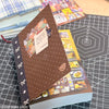 A close-up of a brown sticker book with decorative elements and colorful pages resting on a black grid surface.