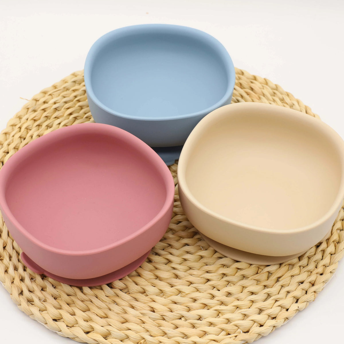 A set of five silicone bowls in pink and beige colors resting on a woven mat.