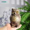 A realistic brown and tan owl garden statue held in a person's hand, set against a green foliage background.