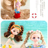 A small doll with brown hair wearing an orange hat and green outfit standing on a sandy beach.