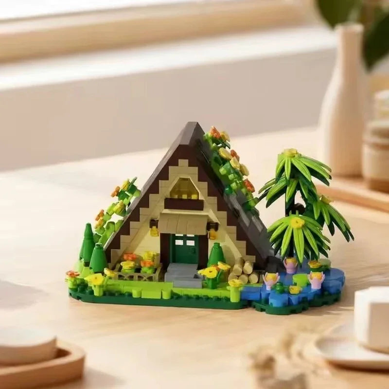 A miniature wooden cabin building set featuring a dark brown roof, green accents, and small plants on a light wood surface.