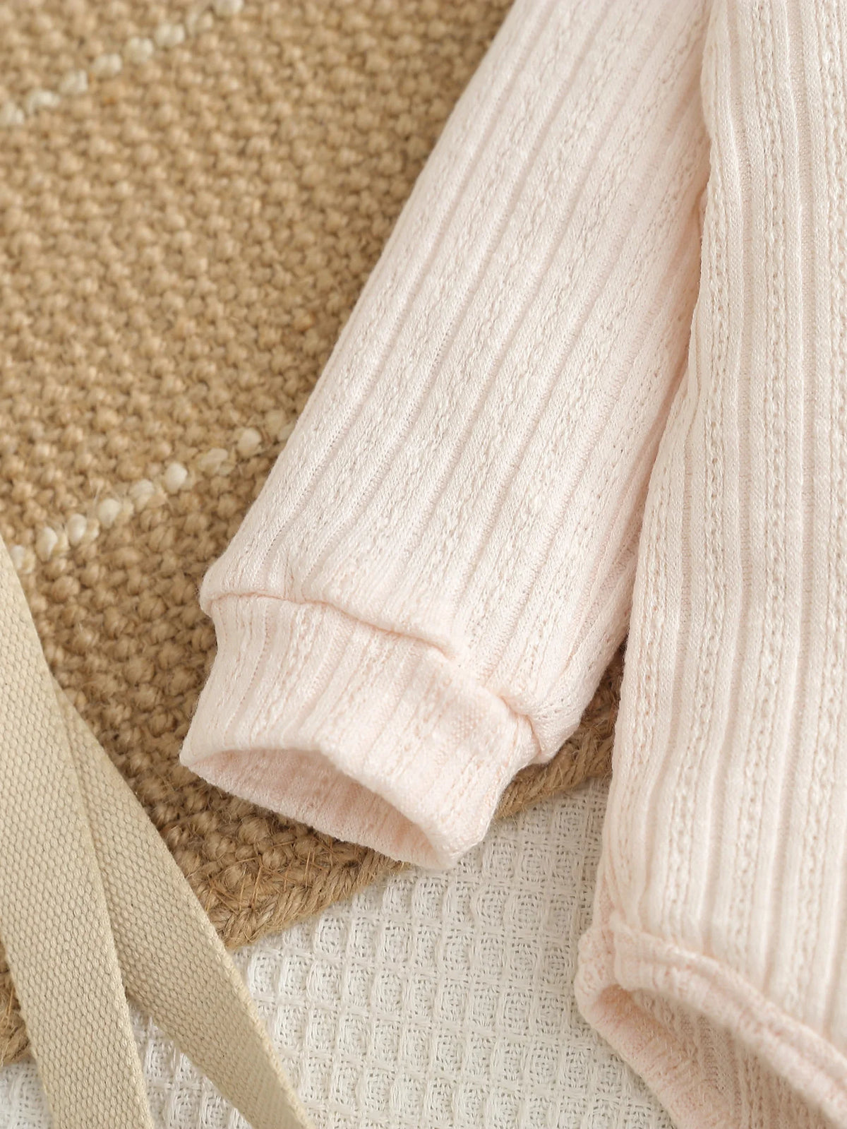 A light pink knitted baby romper and pants set lying on a beige textured surface.