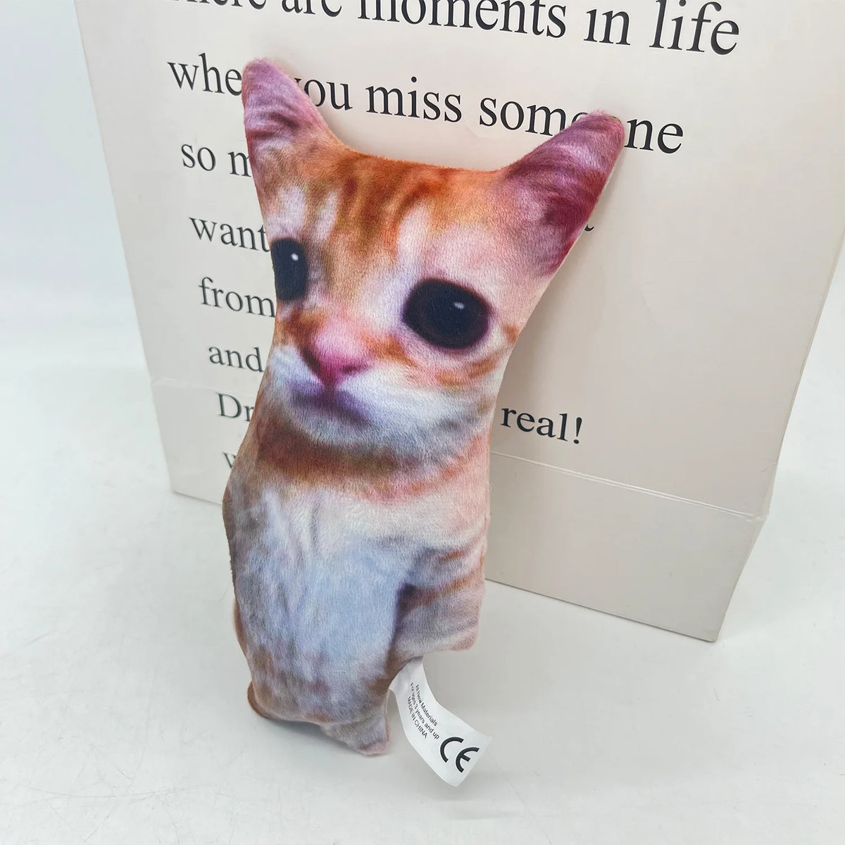 A small, plush cat doll with orange and white fur is displayed on a white box.