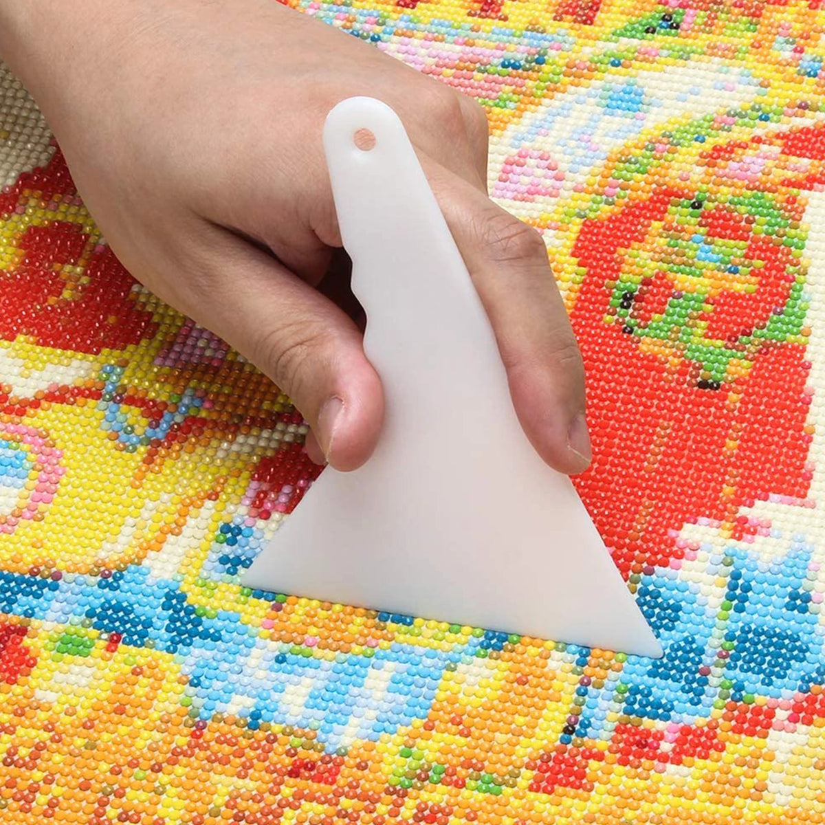 A hand holds a white diamond painting precision tool, showing its shape and size against a colorful diamond painting surface.