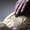A hand uses a chisel to carve intricate patterns into a light-colored wooden object.