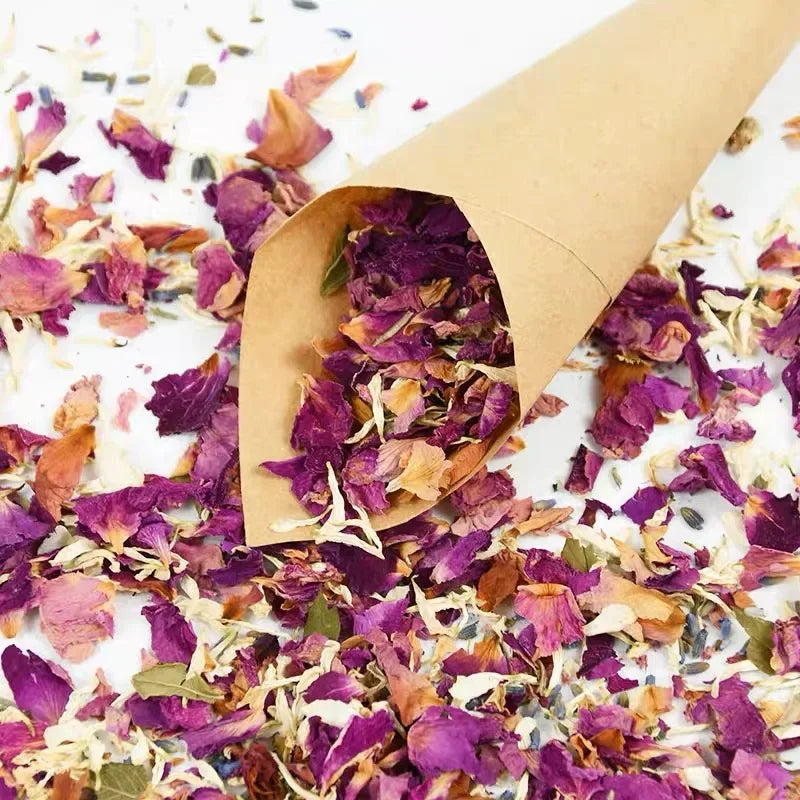 Dried purple and pink rose petals scattered on a white surface inside a brown paper cone.