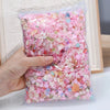 A hand holds a clear plastic bag filled with multicolor resin nail charms in a mix of pink and iridescent colors.