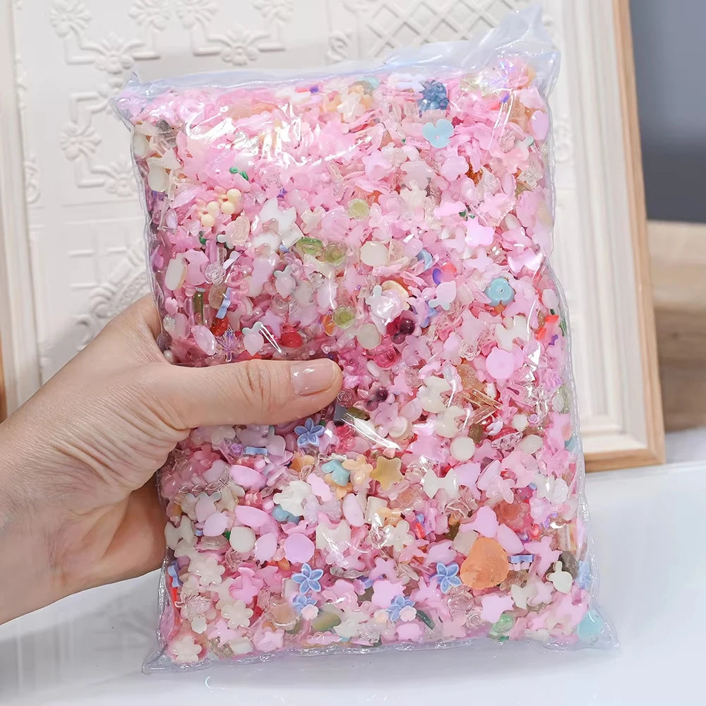 A hand holds a clear plastic bag filled with multicolor resin nail charms in a mix of pink and iridescent colors.
