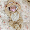 A newborn baby doll wearing a fluffy bear outfit and a pink pacifier is lying on a floral patterned sheet.