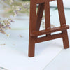 A small, brown wooden artist easel is shown against a white background with some dried flowers.