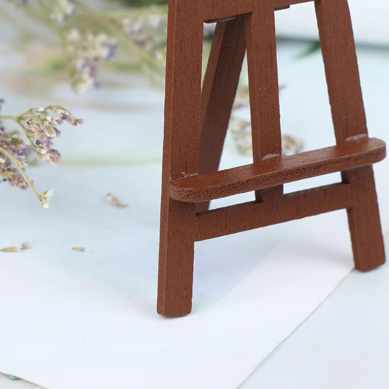 A small, brown wooden artist easel is shown against a white background with some dried flowers.