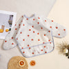A white and pink checkered waterproof long sleeve feeding bib for babies placed on a light background with citrus fruit.