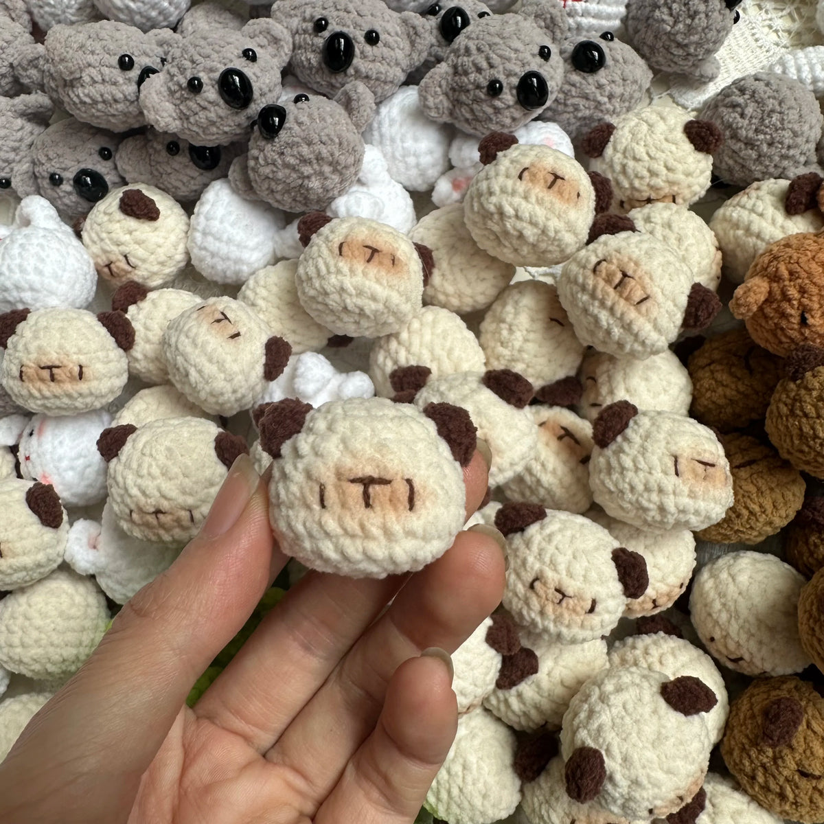 A hand holds a cluster of handcrafted crochet animal keychains, including rabbits and chicks, in various neutral colors.