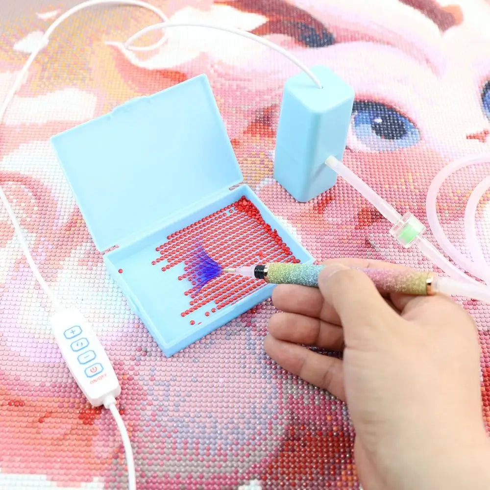 A hand is using an electric diamond painting pen kit with a suction tool to create a design on a pink background.