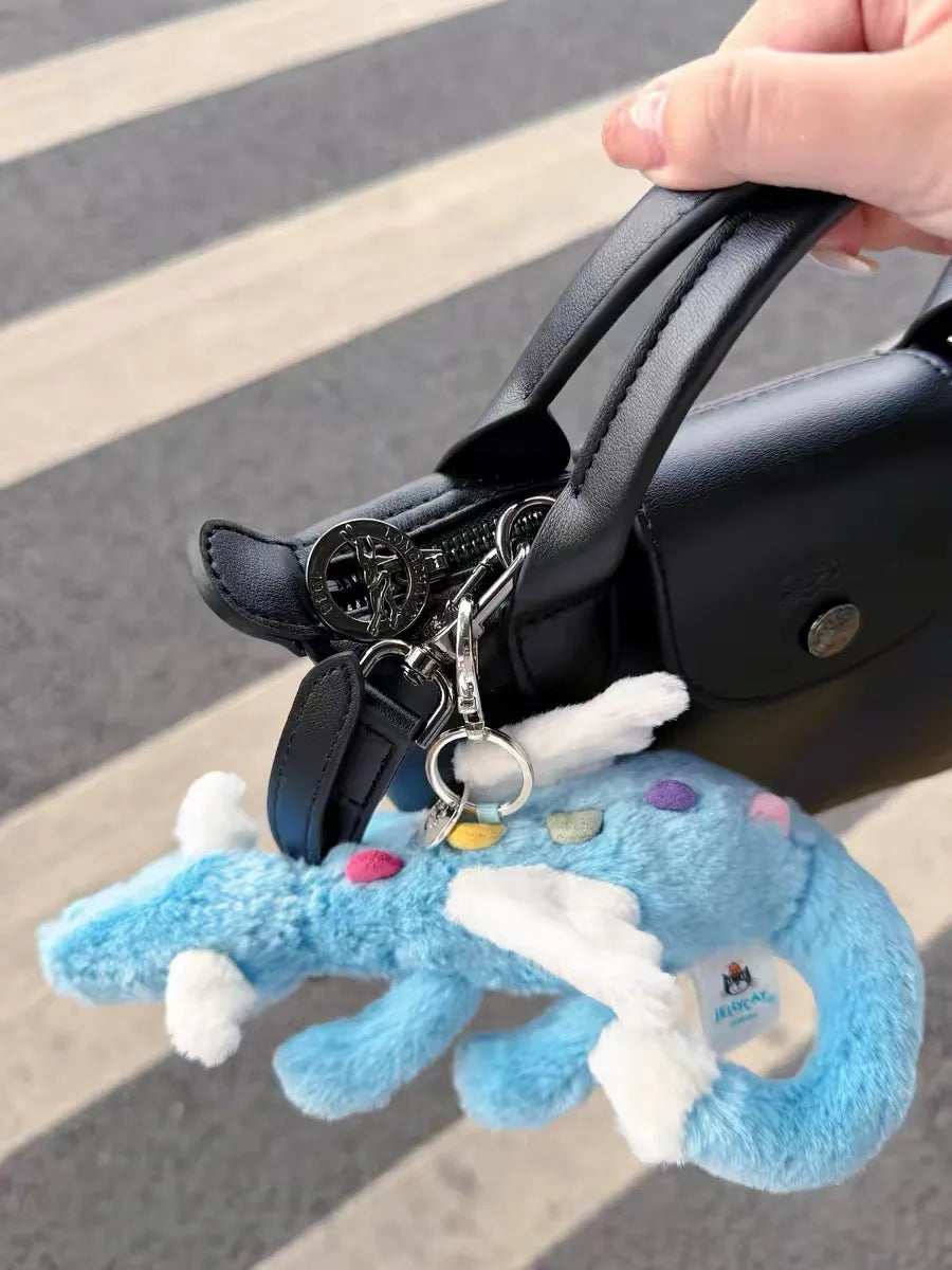 A hand is holding a black bag with a silver charm and a light blue rainbow dragon plush toy.