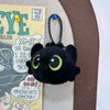 A black plush keychain of Toothless the dragon with large green eyes and a black cord.