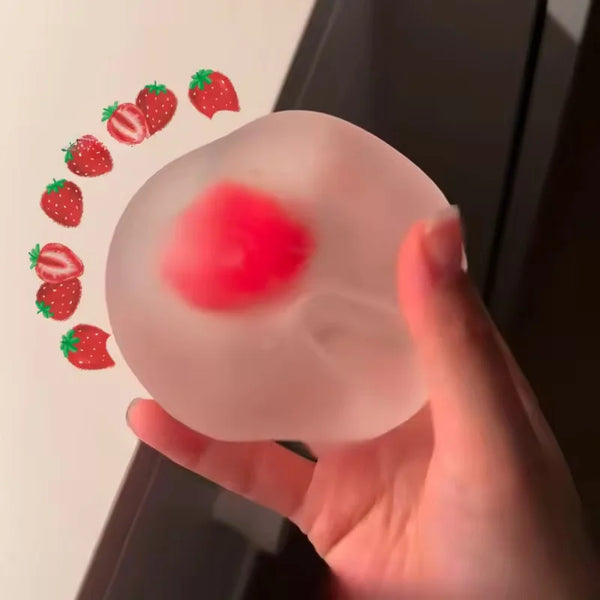 A hand holds a translucent pink squishy stress ball shaped like a strawberry, with several small red strawberry shapes floati