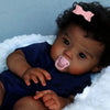 A close-up photo of a Reborn baby doll with hand-rooted curly hair wearing a dark blue outfit and a pink bow, resting on whit