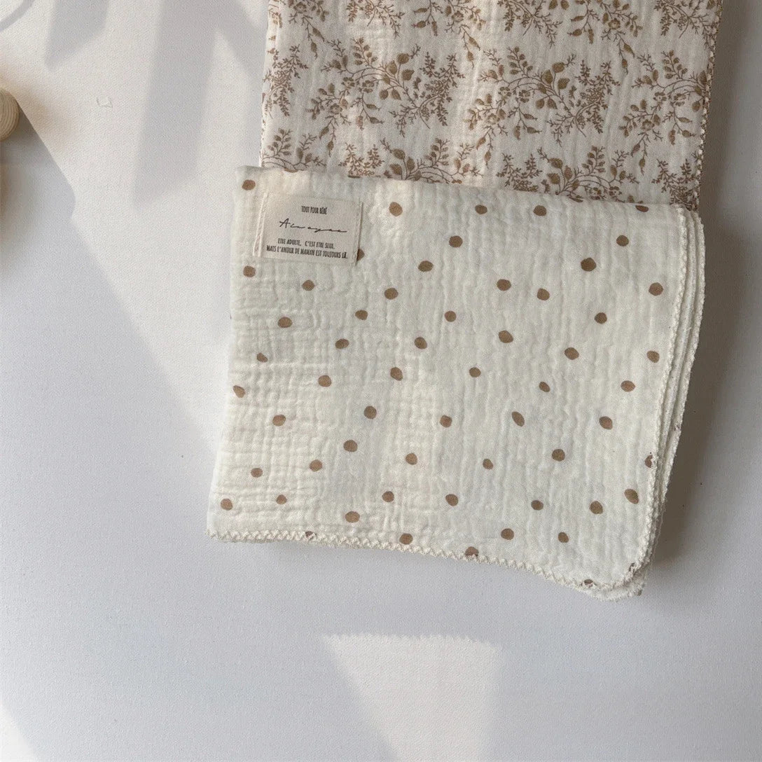 A white muslin swaddle blanket with a floral print is shown folded on a light surface.