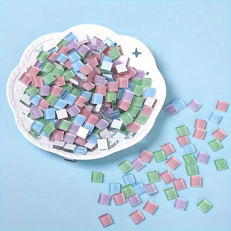 A pile of small, colorful crystal glass mosaic tiles in various shades of green, blue, and pink on a white dish.