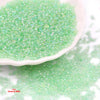 A pile of small, glossy, light green glass seed beads on a white dish and surface.