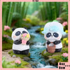 A white and black panda collectible figure with light blue accents sits near a small stream surrounded by green moss and plan