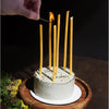 A white cake decorated with birthday candles sits on a wooden platter, with a hand lighting the candles.