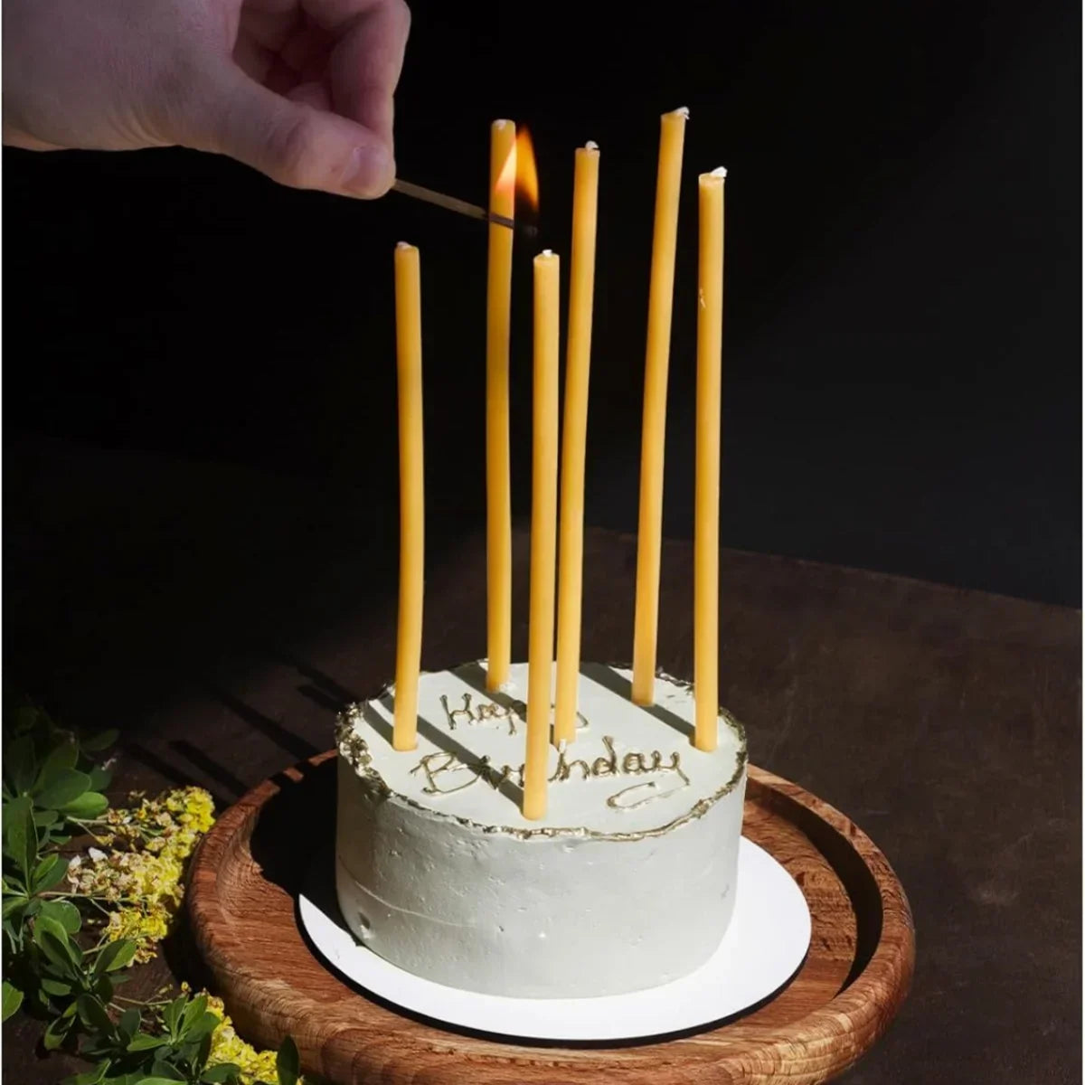 A white cake decorated with birthday candles sits on a wooden platter, with a hand lighting the candles.