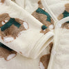 A cream-colored fleece jacket and pants set featuring a brown bear cartoon print with green accents.