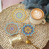 A set of eight colorful mosaic coasters and a holder displayed on a woven mat next to a latte.
