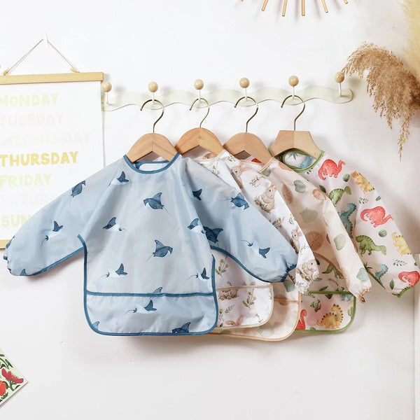 A light blue waterproof long sleeve feeding bib with a pattern of small blue birds hanging on a wooden hanger.