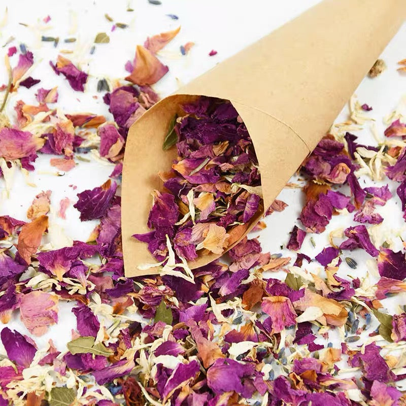 Dried purple and pink rose petals scattered on a white surface inside a brown paper cone.