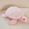 A soft pink sea turtle plush toy with a textured shell and small black eyes resting on a light surface.