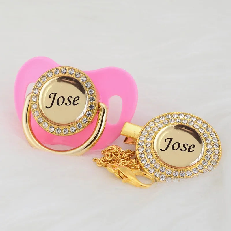 A pink pacifier clip set featuring a gold-toned ring and a crystal charm engraved with the name Jose