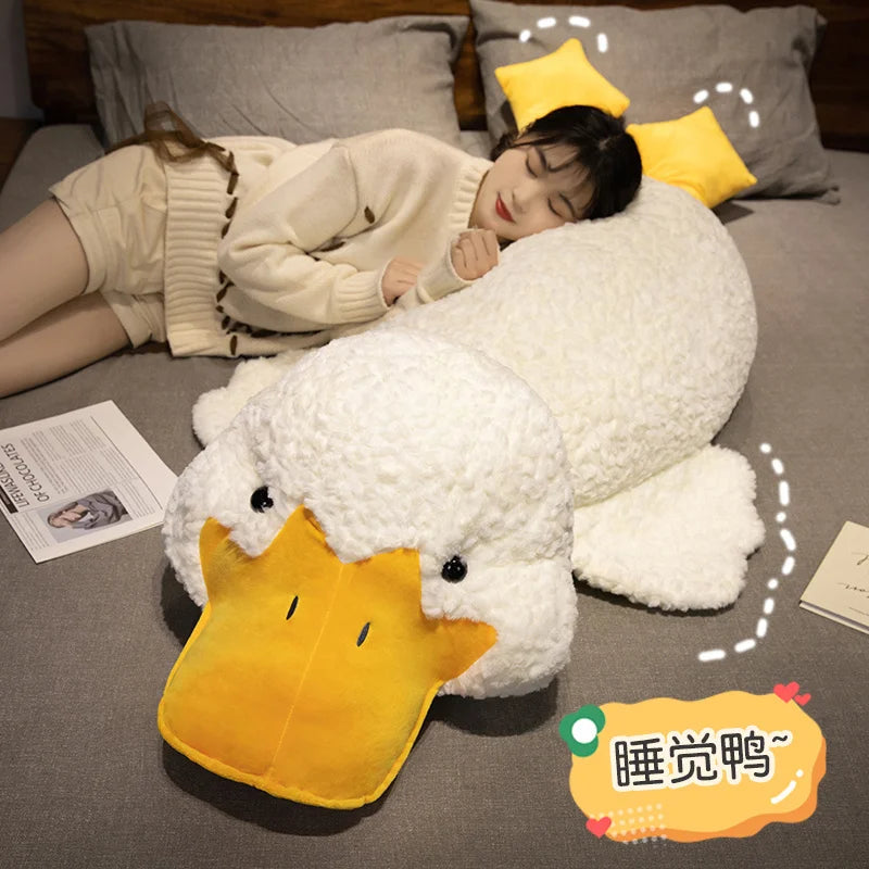 A white plush duck pillow with a yellow beak is resting on a gray cushion next to a person lying down.