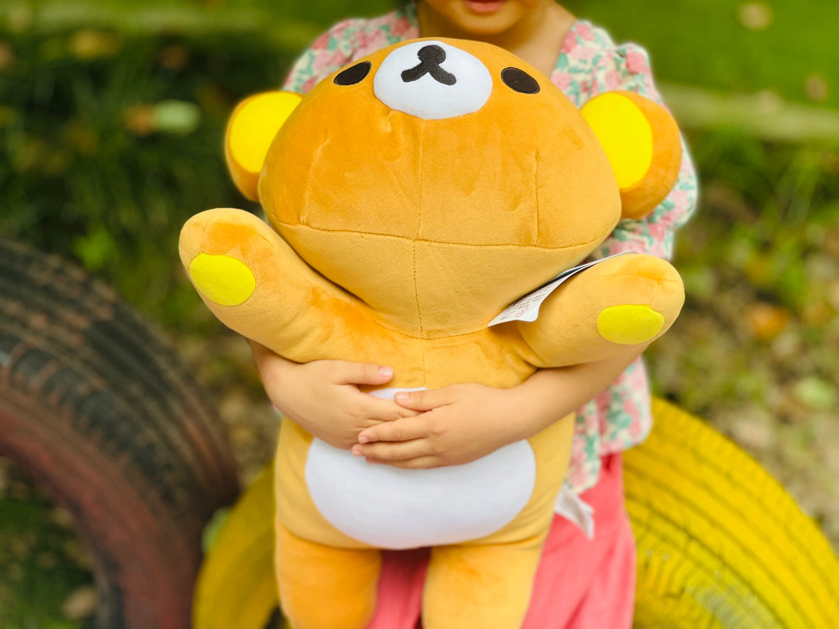 A child is holding a yellow Rilakkuma anime plush bear with large eyes outdoors.