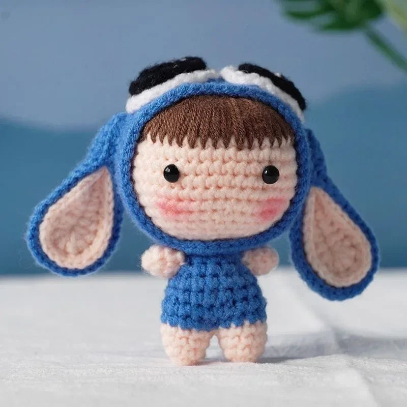 A handmade blue crochet doll with black hair and large ears, sitting on a white surface.