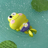 A yellow wind-up swimming frog toy floating on green water with a green leaf visible.