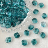 A collection of teal blue geometric glass beads in irregular square shapes scattered on a white surface.