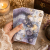 A hand holds a purple and gold oracle card deck with swirling patterns on a rustic surface.