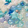 A close-up of various iridescent blue and purple acrylic bow and flower beads for DIY jewelry making.